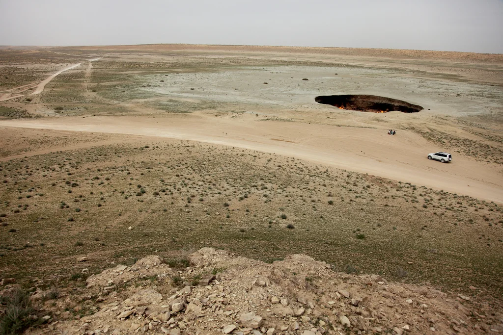 Daytime view from a ridge over the Darvaza gas crater in the Karakum Desert, with a white 4x4 and two visitors near the rim.