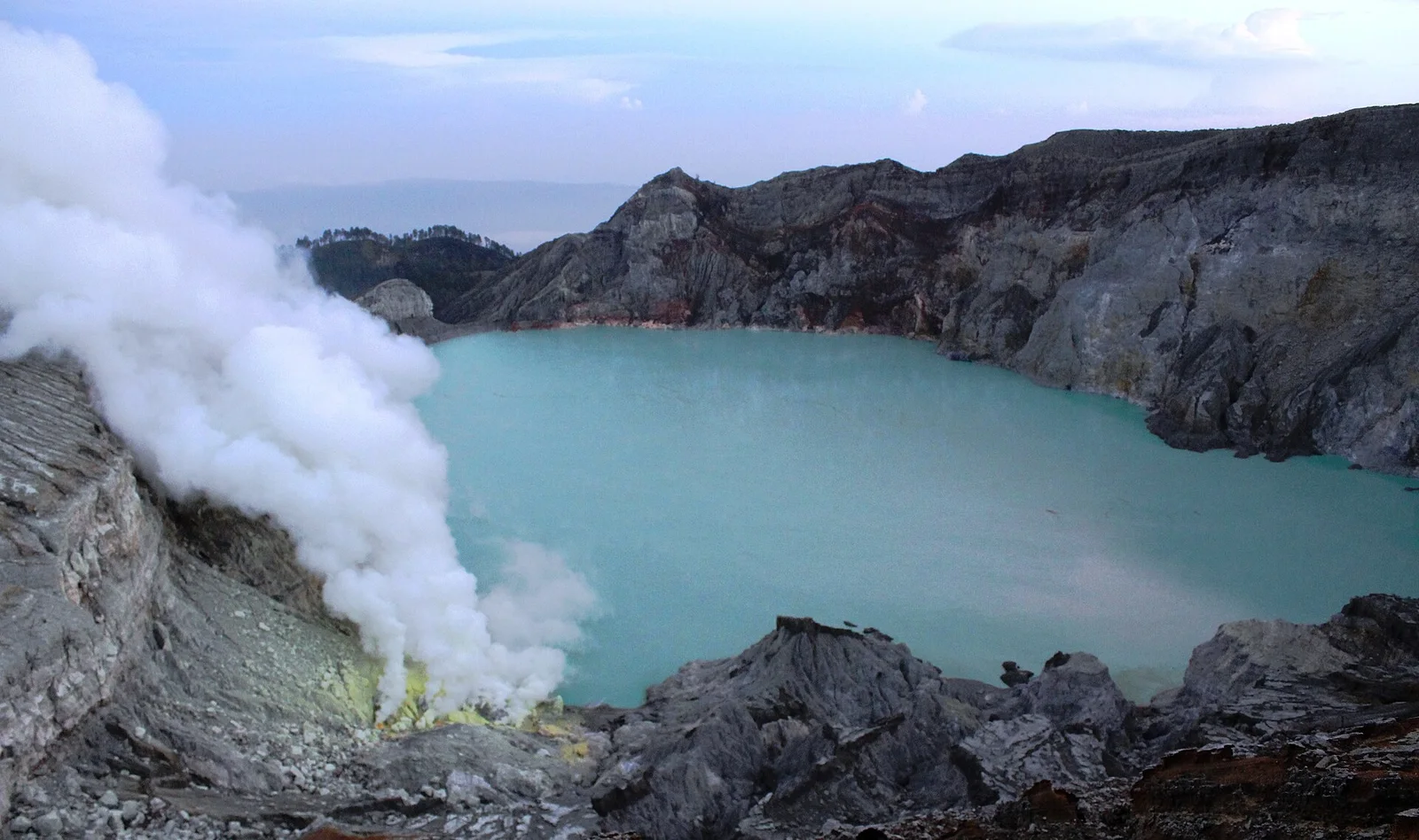 Sulfur fumes billow beside the turquoise acid lake inside Kawah Ijen's crater, ringed by grey volcanic walls.