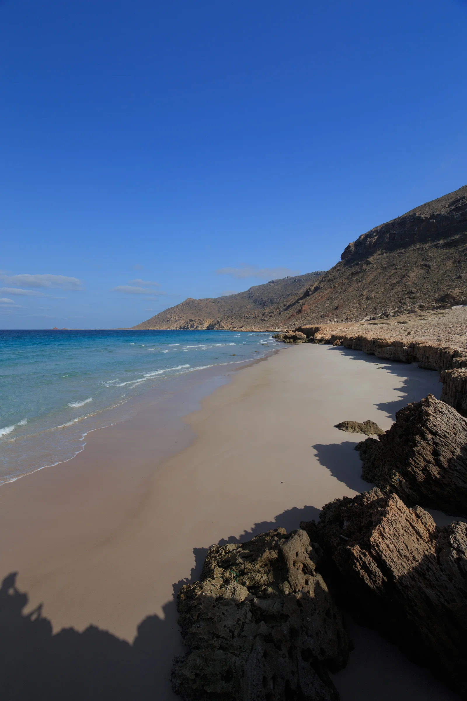 A wide, empty sand beach curves beneath steep brown cliffs beside turquoise water at Delisha, Socotra.